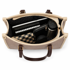 An open beige and brown tote bag shown from above, revealing its interior storage capacity with a black patterned lining, a notebook, a wallet, a tumbler, and a plaid fabric insert on a white background.